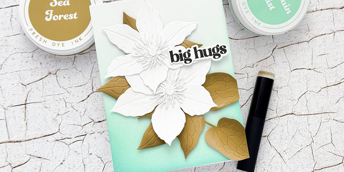 Handmade floral card using a die cut hack to perfectly arrange white Clematis flowers and gold leaves over a teal ink-blended background.