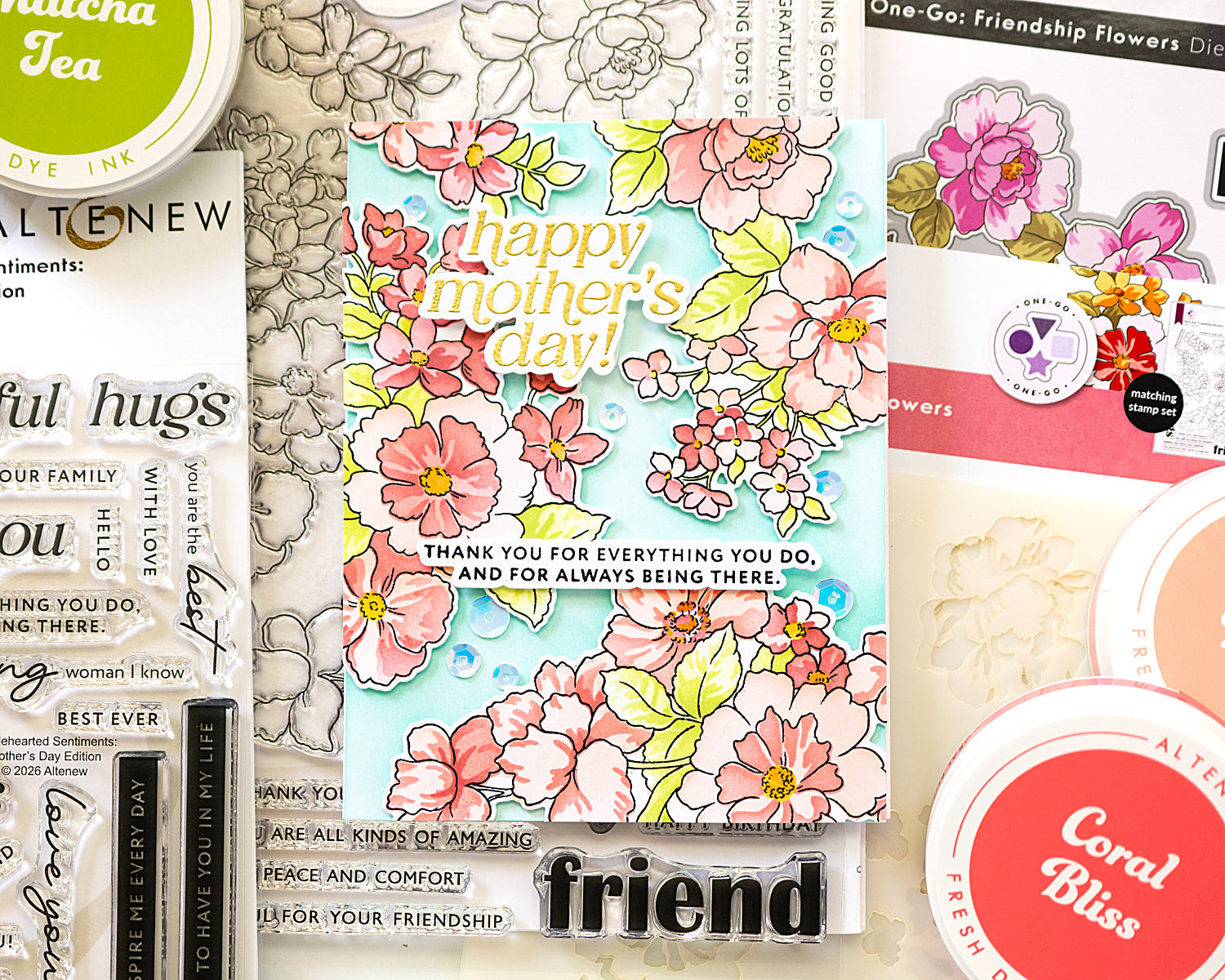 Handmade Mother’s Day card with pastel ink‑blended florals, layered die cuts, and coordinating crafting supplies arranged around the finished design.