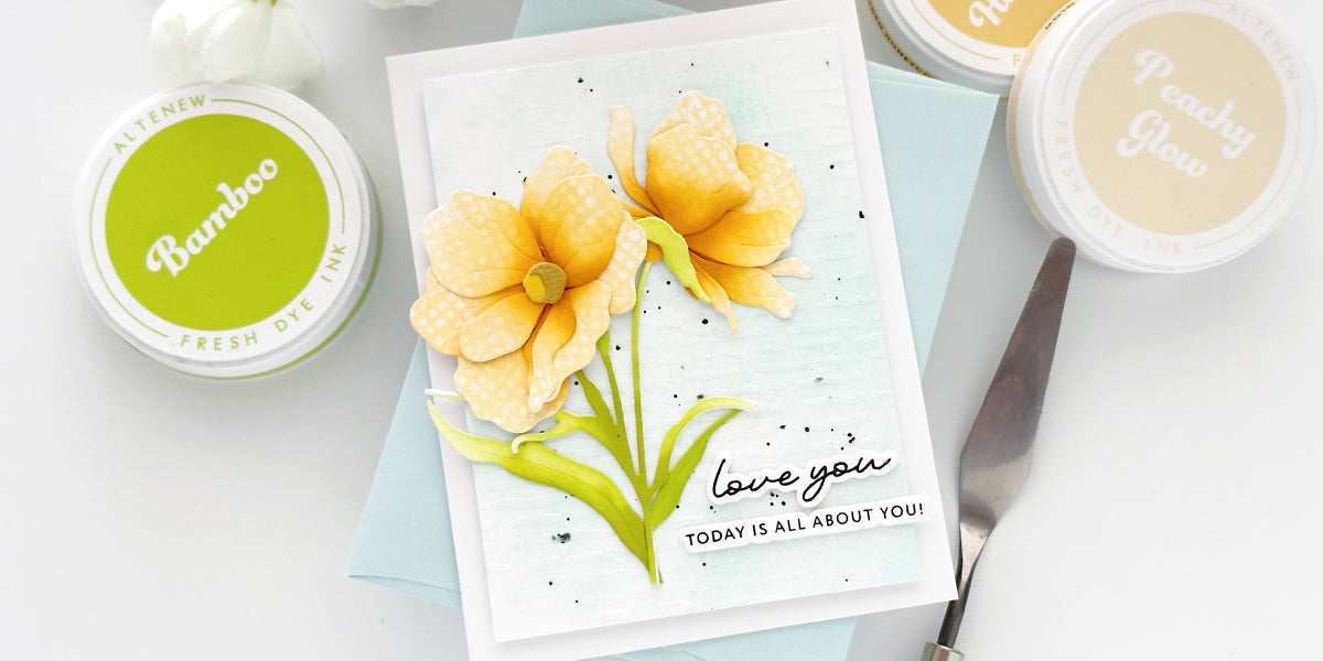 Handmade card featuring two large, yellow-layered die-cut flowers with a white patterned texture on the petals. The flowers have green stems and are set against a pale blue, textured background with black ink splatters. A sentiment at the bottom reads 
